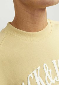 Sweatshirt jaune avec un col rond côtes, présentant un texte brodé blanc. Texture de tissu lisse avec des détails de couture visibles.