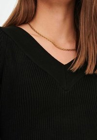 Mujer que lleva un suéter negro de canalé con escote en V y una simple cadena de oro, con el cabello lacio castaño claro sobre un hombro.