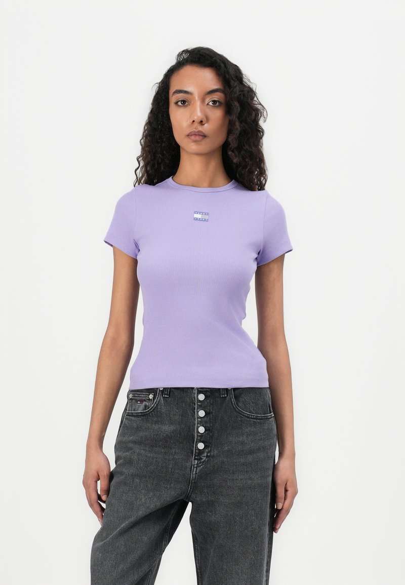 T-shirt lilas à manches courtes et col rond avec une petite étiquette graphique sur la poitrine, associé à un jean taille haute gris.