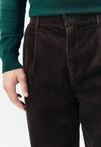Close-up of a person's hand resting near dark brown corduroy pants with a button and green long-sleeve shirt visible.