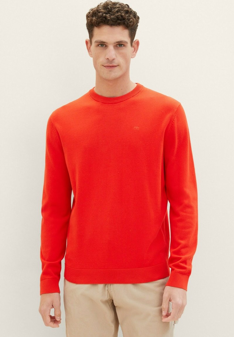 Pull en tricot rouge avec un col rond, des manches longues et des poignets côtelés ; présente un petit logo sur la poitrine.