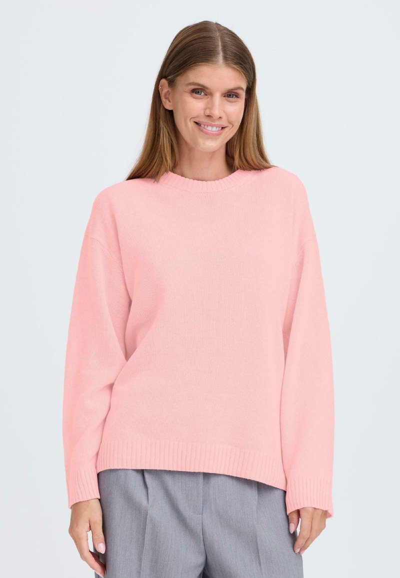 Rosa stickad tröja med rund halsringning, sänkta axlar och ribbad nederkant. Matchad med grå byxor, strukturerad tyg synlig i detalj.