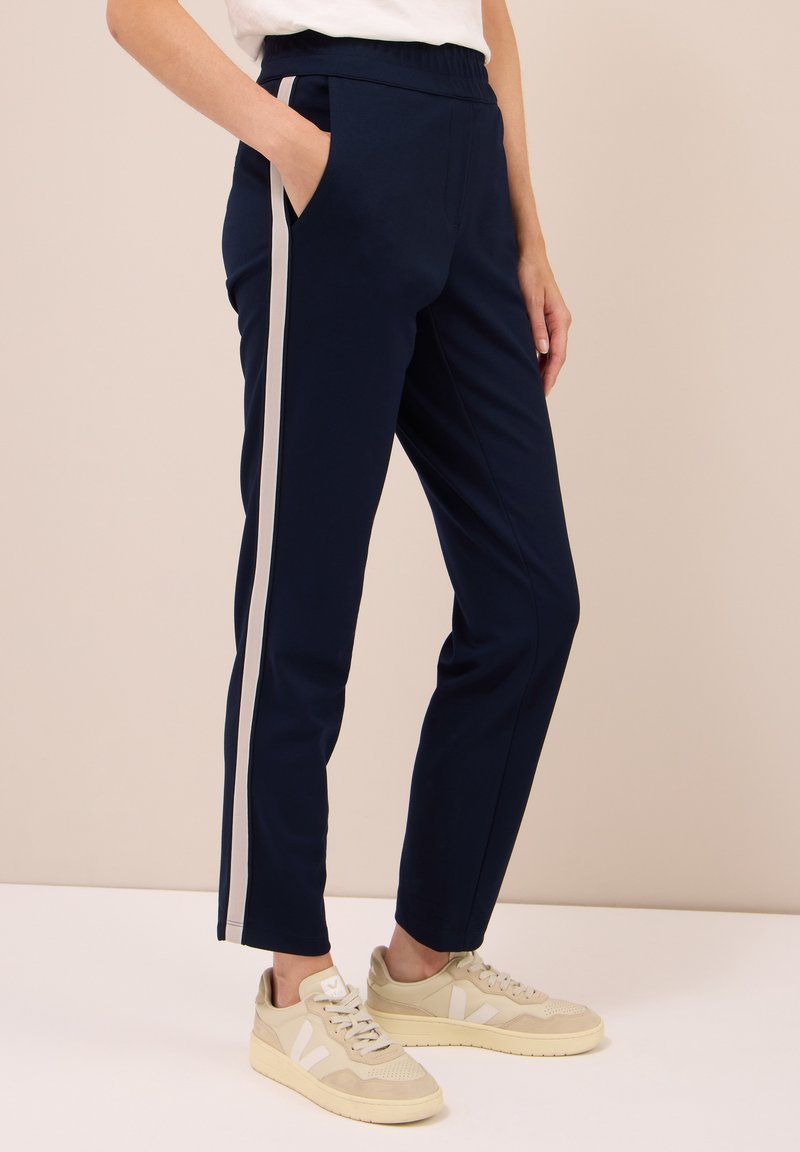 Persona che indossa pantaloni blu navy con strisce bianche sui lati e sneakers beige, in piedi con una mano in tasca su uno sfondo semplice.