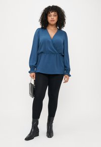 Blouse en teal avec des manches longues et froncées, et une texture subtile, associée à des leggings noirs et des bottes à talons pointues. Tenant un petit sac noir.