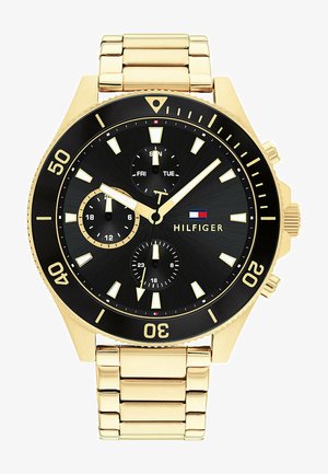 Relógio em metal dourado com mostrador preto, marcadores luminosos e três submostradores. Apresenta uma pulseira texturizada e o logótipo "HILFIGER" ao centro.