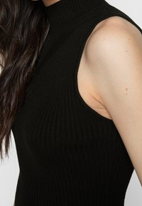 Femme aux longs cheveux foncés portant un haut noir sans manches à col roulé côtelé, sur fond blanc.