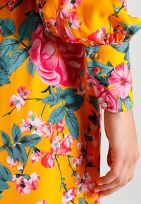 Blommig klänning med en livlig gul bakgrund och rosa samt blå blommor, långa puffärmar och en detaljerad knäppning vid ärmslutet.