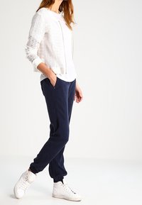 Bílá pletená bomber bunda se zipem v kombinaci s jednoduchým bílým tričkem a námořnickými jogger kalhotami. Doplněno bílými teniskami.