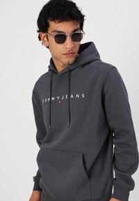 Haut de survêtement gris foncé en tissu doux, doté d'une poche kangourou et du logo blanc "TOMMY JEANS". Il comprend des cordons de serrage ajustables et des poignets côtelés.