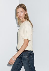 Světle béžové tričko s krátkým rukávem vyrobené z jemného materiálu, spárované s tmavě modrými džíny. Modelka má na sobě stříbrný náramek a stojí proti bílé zdi.