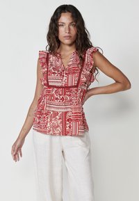 Blusa a fantasia rosso e crema con maniche a volant, scollatura a V e vita arricciata, abbinata a pantaloni leggeri di lino.