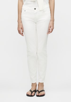 Γυναίκα που φοράει εφαρμοστά λευκά παντελόνια και μαύρα σαγιονάρες, στέκεται μπροστά σε απλό λευκό φόντο.