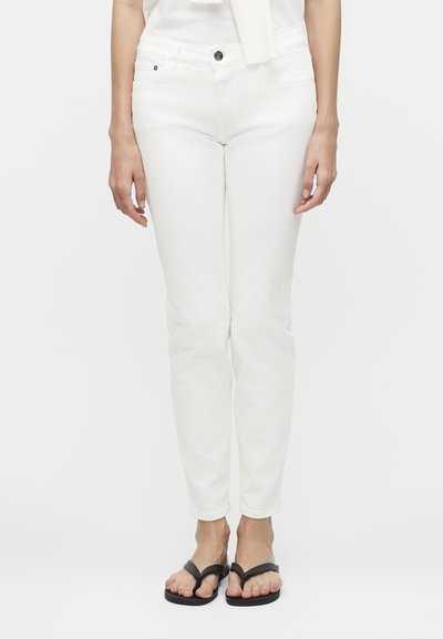Femme portant un pantalon blanc ajusté et des tongs noires, debout devant un fond blanc uni.