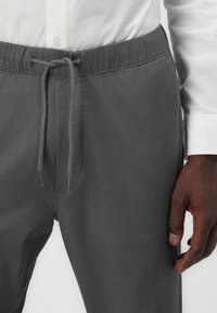 Gray drawstring pants with a textured fabric, elastic waistband, and metal aglets, paired with a white button-up shirt in the background.