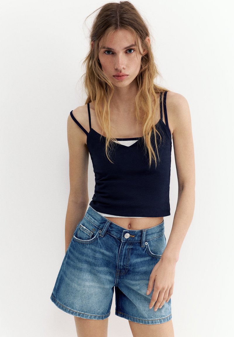 Jeune femme aux longs cheveux blonds portant un haut noir à fines bretelles et un short en jean bleu, debout devant un fond blanc uni.