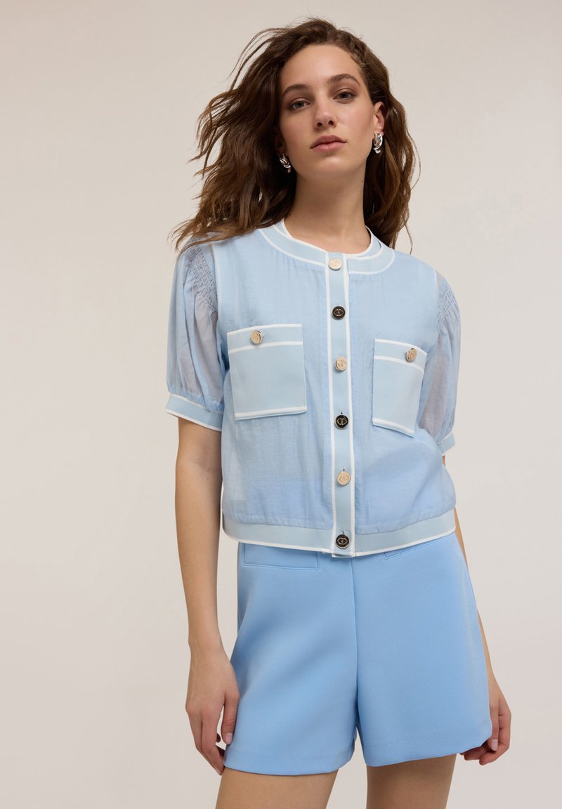Blusa azzurra a maniche corte con colletto tondo, dotata di due tasche frontali e rifiniture bianche in contrasto; bottoni lungo la parte frontale.