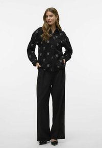 Zwarte button-up blouse met witte strikpatronen, lange mouwen, relaxte pasvorm, gecombineerd met zwarte wijde broek enpuntige schoenen.