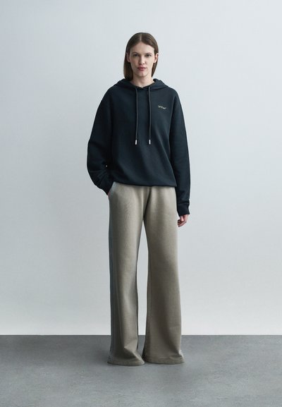 Γυναίκα όρθια, φοράει μαύρο φούτερ με κορδόνια και μπεζ παντελόνα με φαρδιά μπατζάκια, μπροστά σε απλό ανοιχτό γκρι φόντο.