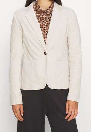 Kvinde iført en beige enkelknaps-blazer over en brun mønstret bluse og sorte bukser, stående mod en ensfarvet baggrund.