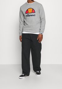 Sweat-shirt gris avec logo coloré, associé à un pantalon noir ample et des baskets noires à semelles blanches. Tissu texturé visible.