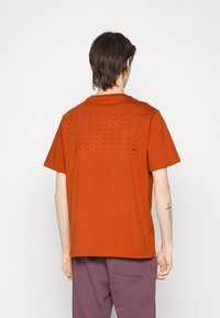 Katoenen oranje T-shirt met korte mouwen en een ronde hals. De achterkant is voorzien van gelijkmatig verdeelde kleine decoratieve gaatjes en een logo.