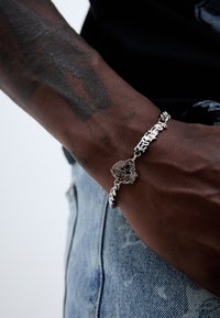 Hõbe käevõru ketidisainiga, millel on detailne ripats. Kantud randmel, kus on nähtavad tätoveeringud, sinise teksakangast taustal.