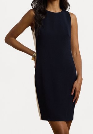 Vrouw draagt een mouwloze, knielange zwarte jurk met witte zijpanelen, staat met één hand op haar heup en de andere ontspannen langs haar zij.