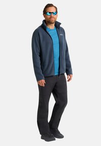 Uomo che indossa una giacca in pile blu, una camicia teal, pantaloni neri, scarpe nere e occhiali da sole blu riflettenti, in piedi contro uno sfondo semplice.