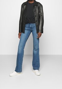 Person iført sort læderjakke, sort knyttet skjorte, blå jeans og hvide sneakers står foran en ensfarvet hvid baggrund.
