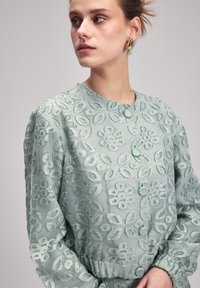 Mintgrön blus med blommig broderi, rund halsringning och gröna knappar. Långa ärmar med elastiska manschetter, strukturerad tyg och intrikat mönster.