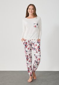 Λευκό πουκάμισο με μακριά μανίκια και λουλουδάτη λεπτομέρεια στην τσέπη, συνδυασμένο με παντελόνια με λουλουδάτο σχέδιο σε κόκκινο και μπλε χρώμα σε ανοιχτό φόντο.