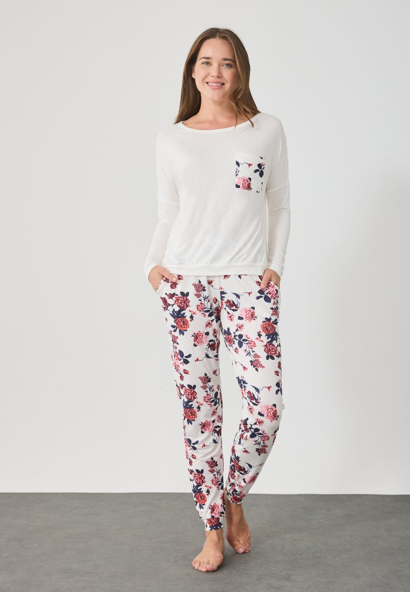 Λευκό πουκάμισο με μακριά μανίκια και λουλουδάτη λεπτομέρεια στην τσέπη, συνδυασμένο με παντελόνια με λουλουδάτο σχέδιο σε κόκκινο και μπλε χρώμα σε ανοιχτό φόντο.