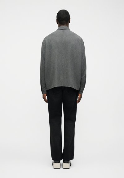 Άνδρας που στέκεται πλάτη φορά γκρι μακρυμάνικο πουκάμισο, μαύρο παντελόνι και παπούτσια με τετράγωνη φτέρνα σε μαύρο και λευκό σε απλό φόντο.