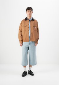 Joven con una chaqueta marrón de cuello negro, camisa de rayas azules, jeans cortos anchos de un azul claro, calcetines blancos y mocasines negros, de pie sobre un fondo blanco.