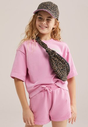 Naeratav tüdruk, kes kannab roosat lühikeste varrukatega särki ja lühikesi pükse, leopardimustrilise nokamütsi ja sobiva ristihoidmisega kotti, silmapilguga ühe silmaga.