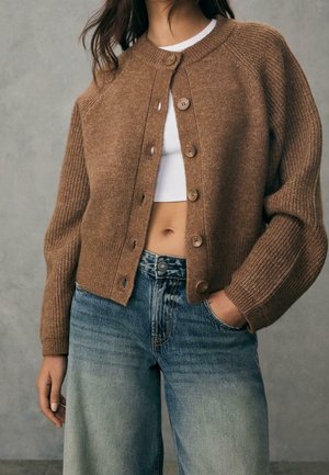 Vrouw draagt een bruine gebreide cardigan met knopen over een wit crop topje en vervaagde blauwe jeans, met één hand in de zak, voor een effen achtergrond.