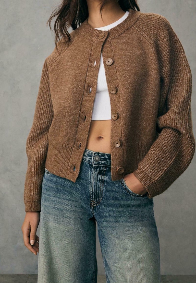 Femme portant un cardigan marron boutonné par-dessus un crop top blanc avec un jean bleu délavé, une main dans la poche, sur un fond uni.