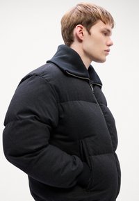 Black puffer jacket with a high collar, featuring a zippered front and side pockets. The texture is smooth with a padded appearance.