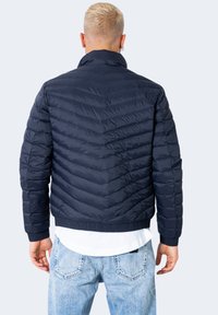 Giubbotto imbottito blu navy con cuciture a chevron, colletto alto e polsini elastici. La texture è liscia, con orlo a coste e dettagli con logo.