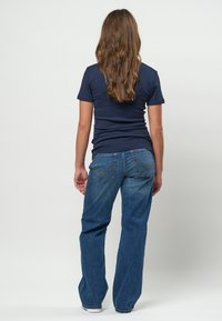 Kvinde med langt brunt hår iført en navyblå kortærmet skjorte og blå jeans, stående med ryggen mod en ensartet hvid baggrund.