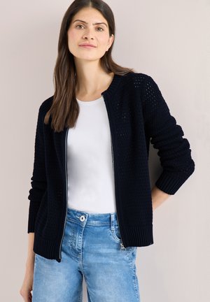 Donna con capelli castani lisci che indossa un cardigan nero con zip sopra una camicia bianca e jeans blu, in piedi contro una parete chiara e semplice.