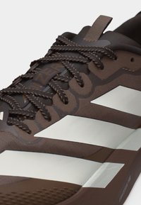 Κοντινή λήψη καφέ αθλητικού παπουτσιού με μαύρες και μπεζ λεπτομέρειες, υφασμάτινα κορδόνια, επάνω μέρος από ύφασμα και θηλιά έλξης στη γλώσσα.