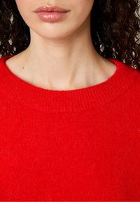 Raudonas megztinis su giliu kaklu. Jo tekstūra atrodo minkšta ir pūkuota, rodanti vientisą spalvą be raštų ar puošmenų.