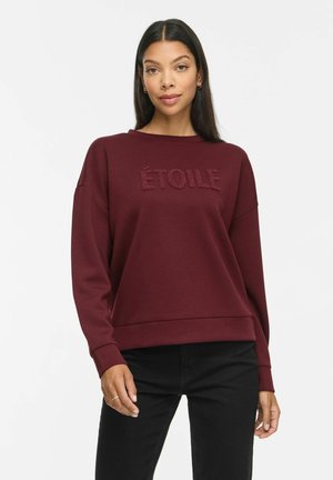 Kvinde med langt mørkt hår iført en burgundy sweatshirt med teksten "ÉTOILE" og sorte bukser, stående mod en ensfarvet lys baggrund.