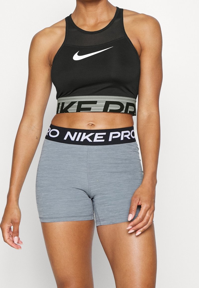 Femme athlétique portant un soutien-gorge de sport noir Nike et un short ajusté gris Nike Pro, debout contre un fond blanc.