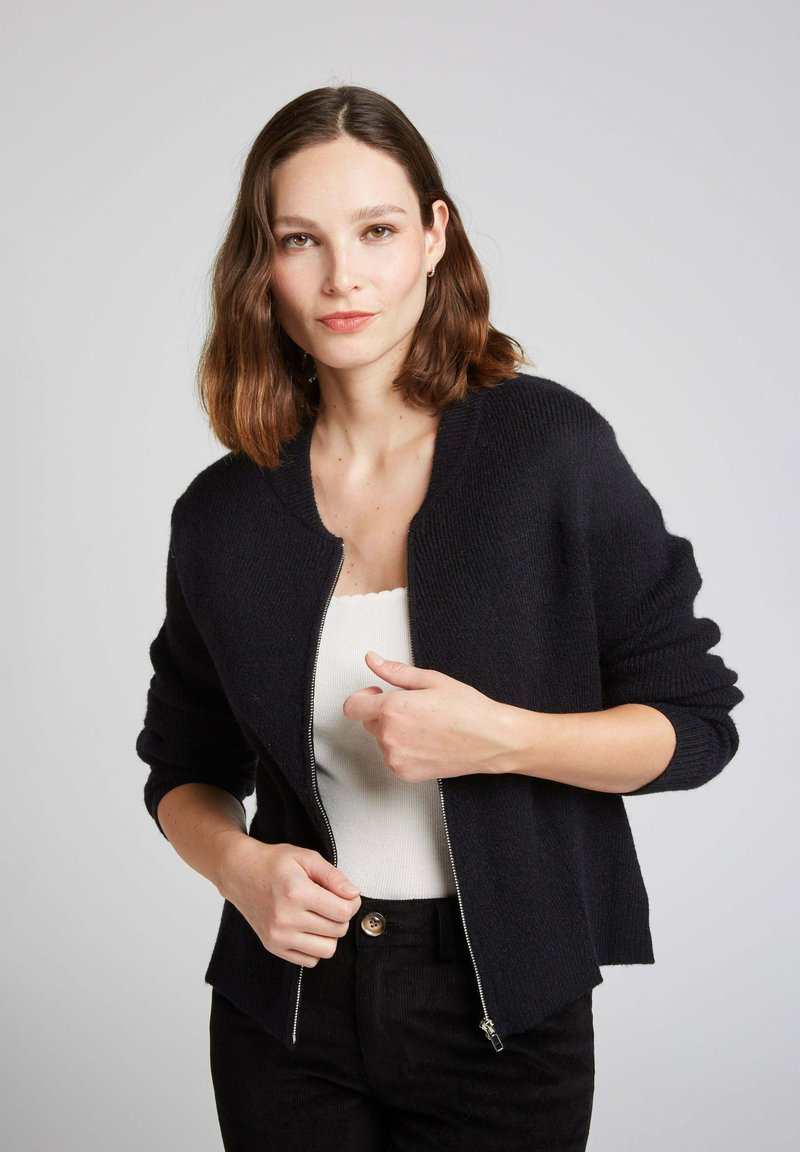 Cardigan noir en tricot avec un col montant, fermeture éclair et poignets côtelés. Porté sur un haut blanc, avec une texture douce et une coupe ajustée.