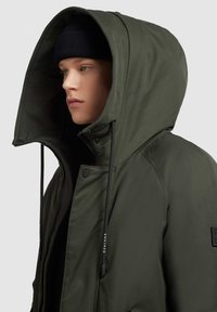 Grøn hættejakke med lynlåslukning, snore og et tekstureret stof. Bæres med en sort strikket hue.