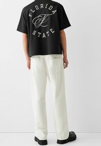 Czarny t-shirt z krótkim rękawem z białym napisem "FLORIDA STATE" na plecach, zestawiony z białymi luźnymi jeansami i czarnymi butami.