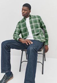 Green plaid shirt with white accents, buttoned-up, worn over a white t-shirt. Paired with dark denim jeans and black shoes. Sitting on a chair.