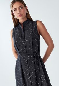 Czarna bezrękawnikowa sukienka w białe kropki, zapinana na guziki z przodu, z kołnierzykiem mandarynkowym i materiałowym paskiem w talii. Gładka, lejąca się faktura.